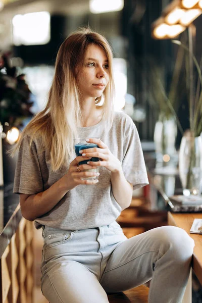 Ein gut aussehendes, schlankes blondes Mädchen, lässig gekleidet, sitzt auf dem Stuhl und hält eine Tasse Kaffee in einem gemütlichen Café. — Stockfoto