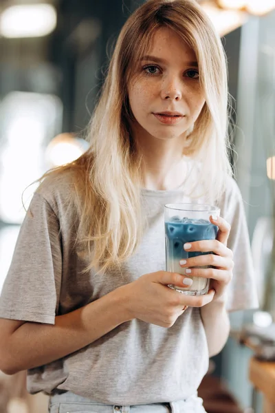 Μια εμφανίσιμη λεπτή ξανθιά κοπέλα, φορούν casual στυλ, κάθεται στην καρέκλα και κατέχει ένα φλυτζάνι του καφέ σε ένα άνετο κατάστημα καφέ. — Φωτογραφία Αρχείου