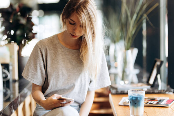 A cute thin blonde girl,dressed in casual style,drinks coffee and looks at her phone in a coffee shop.