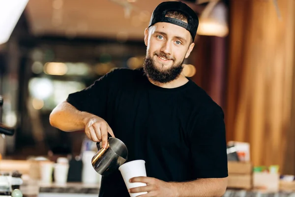 Красивий молодий чоловік з бородою, одягнений у казуальний стиль, додає молоко до кави в сучасній кав'ярні . — стокове фото