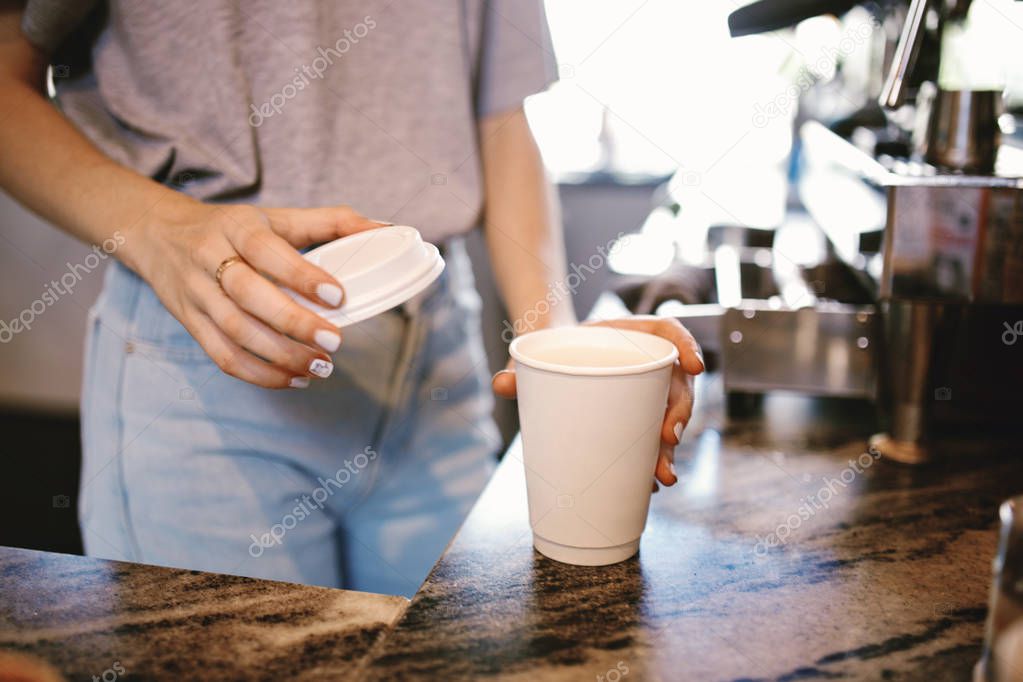 A slim young beautiful girl,dressed in casual outfit, gently puts the lid on the coffeein a cozy coffee shop.