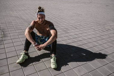 Kafa bandı ve dövmeler ile çıplak bir gövde ile genç atletik erkek siyah tayt giymiş ve mavi şort oturur üzerinde kaldırım döşeme sıcak güneşli bir günde