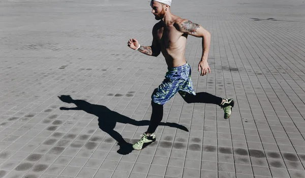 Homem atlético com um torso nu com tatuagens e headband em sua cabeça vestida com as leggings pretas e shorts azuis corre em lajes de pavimentação em um dia ensolarado quente — Fotografia de Stock