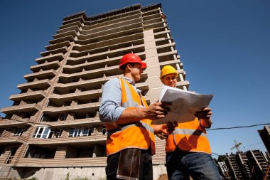 İki yapısal mühendisleri gömlekler, turuncu iş yelek giymiş ve kask multistorey bir binanın arka planı inşaat dokümantasyon keşfetmek