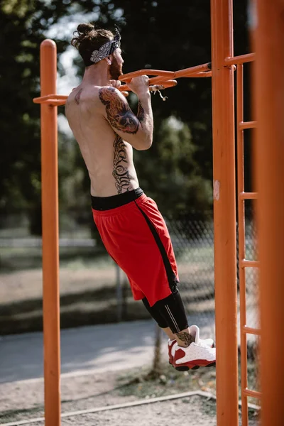 Young athletic man in headband with naked torso dressed in black leggings and red shorts pulls up on the horizontal bar on the sports ground outside on a sunny day