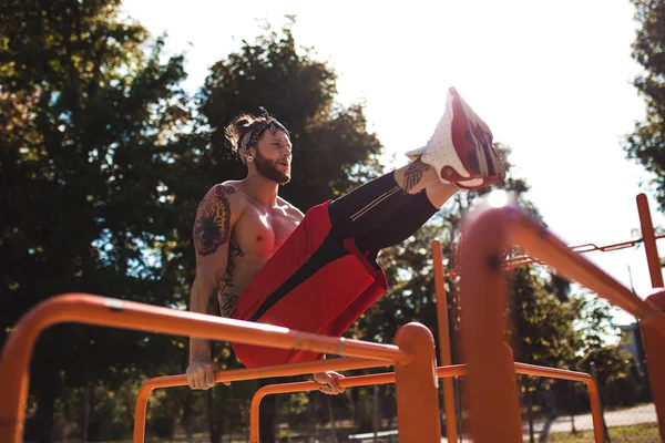 Jeune homme en bandeau avec torse nu vêtu de leggings noirs et short rouge faisant des exercices de presse sur la barre horizontale sur le terrain de sport à l'extérieur par une journée ensoleillée — Photo