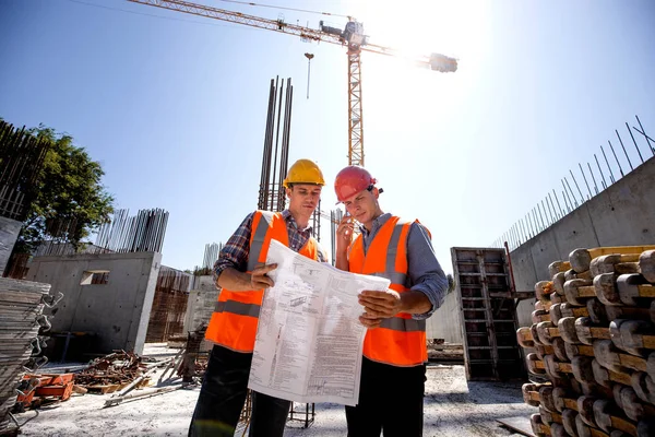 Structural engineer and construction manager in orange work vests and hard helmets discuss documentation and talk by phone on the open air building site next to the crane — Stock Photo, Image