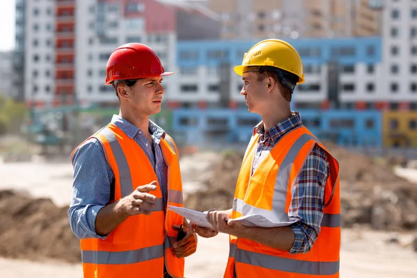 Två män klädda i skjortor, orange arbete västar och hjälmar diskutera dokumentation på byggnadsplatsen mot bakgrund av en flera våningar byggnad — Stockfoto