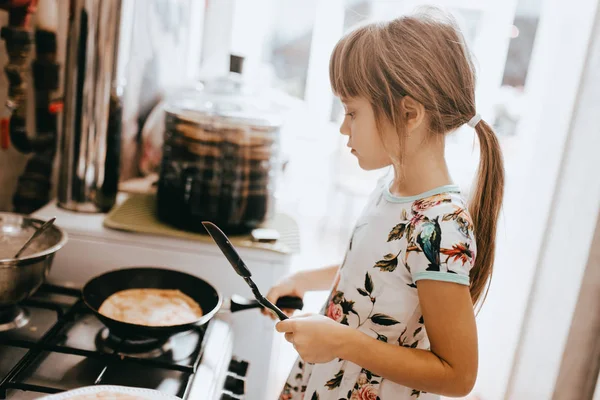 Little girl is cooking pancakes for the breakfast in the little cozy kitchen — Stock Photo, Image