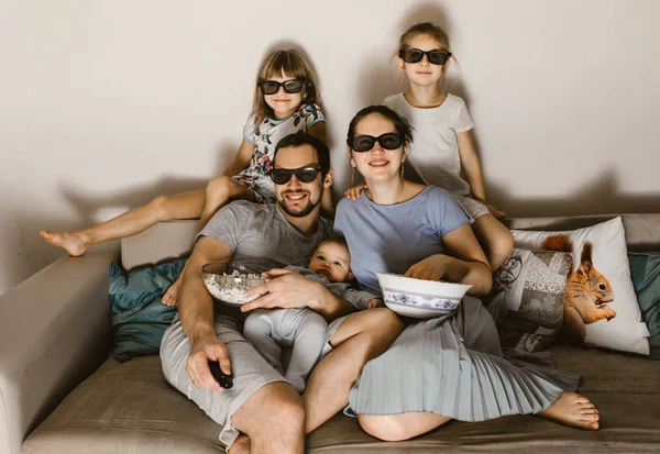 Щасливий батько сім'ї з дитиною на руках, мати і дві дочки в спеціальних окулярах дивитися телевізор і їсти попкорн, сидячи на дивані — стокове фото