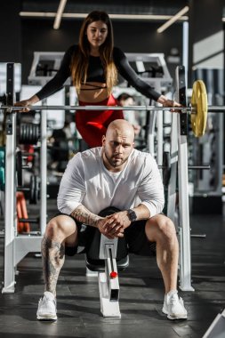 Acımasız adam atletik spor bankta oturur ve ince kız arkasından modern spor salonunda Spor donatımı yanında duruyor