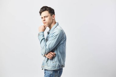 Stüdyoda beyaz arka plan üzerinde beyaz bir t-shirt ve kot ceket giymiş düşünceli esmer adam duruyor