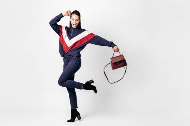 Funny girl dressed in sporty blue suit with a red and white print on a sweatshirt and heels poses with bag in her hands on the white background in the studio clipart