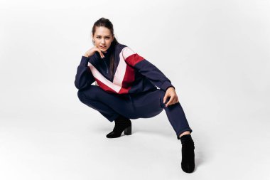 Nice girl dressed in sporty blue suit with a red and white print on a sweatshirt and heels poses squatting on the white background in the studio clipart