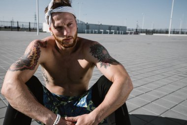 Kafa bandı ve dövmeler ile çıplak bir gövde ile genç atletik erkek siyah tayt giymiş ve mavi şort oturur üzerinde kaldırım döşeme sıcak güneşli bir günde