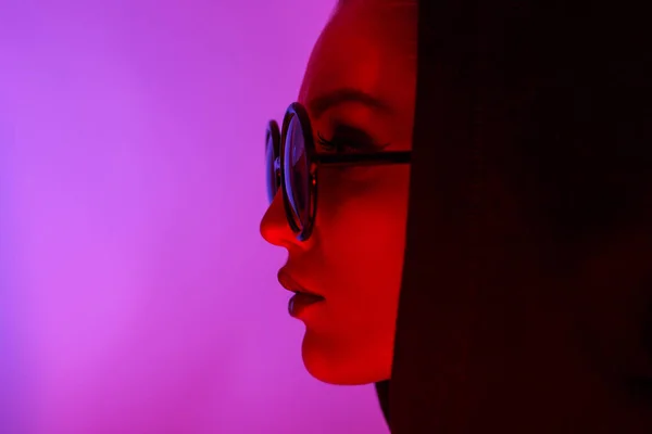 Portrait of fashion young girl in a black sweater with a hood and round sunglasses in red and blue neon light in the studio — Stock Photo, Image