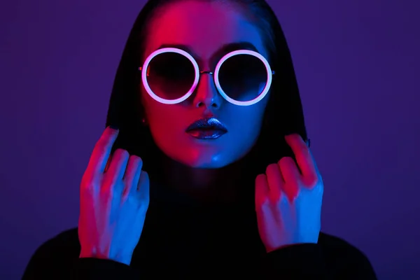 Modemädchen im schwarzen Pullover mit Kapuze und runder Sonnenbrille in rotem und blauem Neonlicht im Atelier — Stockfoto