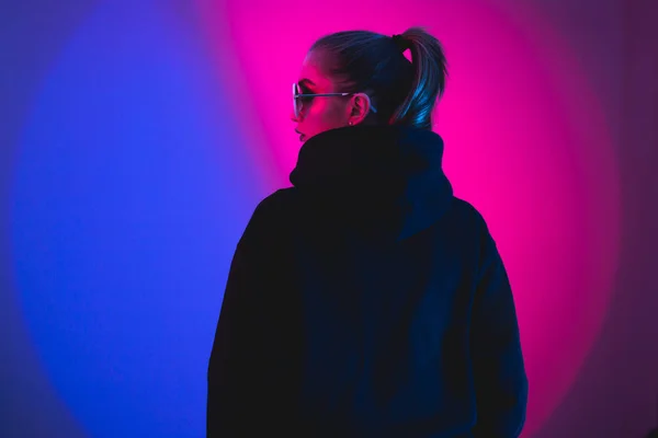 Modemädchen im schwarzen Pullover mit Kapuze und runder Sonnenbrille in rotem und blauem Neonlicht im Atelier — Stockfoto