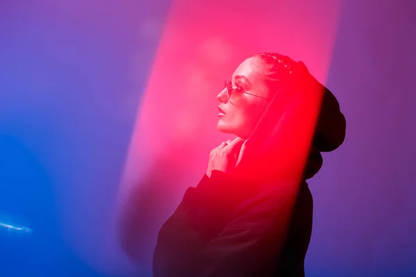 Portrét, fashion Girl v černý svetr s kapucí a sluneční brýle kulatý tvar v neonové světlo v ateliéru — Stock fotografie