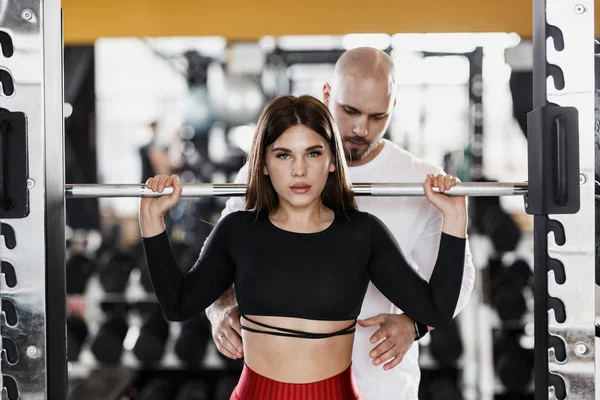 Slender beautiful girl is doing back squats and strong athletic man insures her in the modern gym
