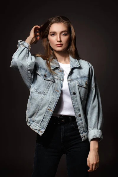 Чудова брюнетка дівчина з довгим волоссям, одягнена в джинсову куртку і джинсову позу, що стоїть на темному тлі в студії — стокове фото