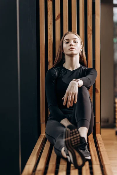 Menina magro bonita vestida em um sportswear preto está sentado no parapeito da janela de madeira no ginásio moderno — Fotografia de Stock