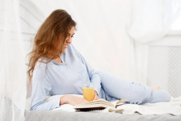 Jolie fille brune vêtue du pyjama bleu clair repose sur le lit avec un verre de jus frais et lit le livre dans la salle de lumière confortable — Photo