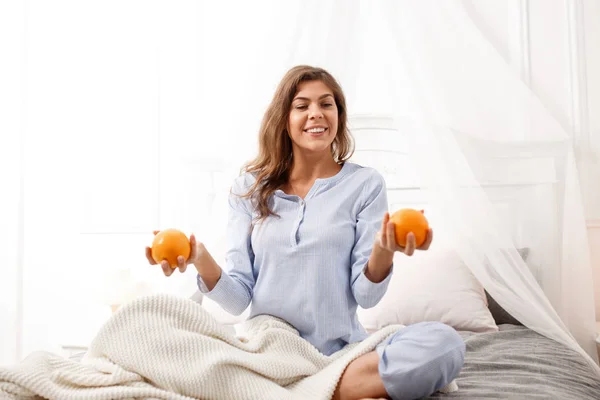 Усміхнена брюнетка дівчина в світло-блакитній піжамі сидить на ліжку з апельсинами в руках на сірому листі з бежевою ковдрою в спальні — стокове фото