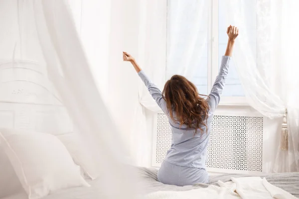 Брюнетка в светло-голубой пижаме вытягивает руки, сидя на кровати с балдахином рядом с окном в уютной спальне. — стоковое фото