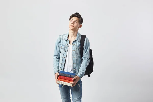 Jovem de cabelos escuros com mochila preta em seu ombro vestida com uma camiseta branca, jeans e uma jaqueta de ganga segura livros em suas mãos no fundo branco no estúdio — Fotografia de Stock