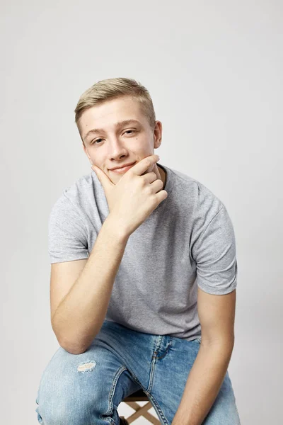 Biondo ragazzo vestito con un bianco t-shirt stand su il bianco sfondo in il studio tenendo il suo mano su il mento — Foto Stock