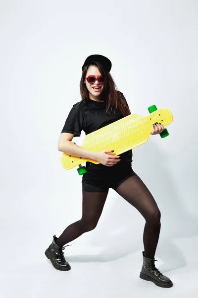 黒のトップ、ショーツ、タイツ、キャップと赤サングラス黄色のスケート ボードとの面白い黒髪少女がスタジオで白い背景にポーズをとってください。 — ストック写真