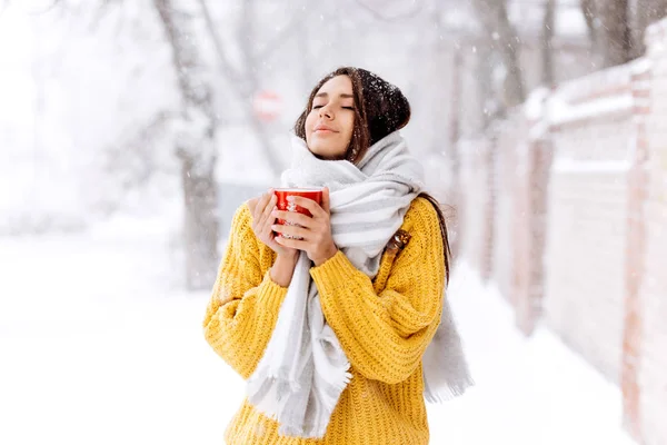 Симпатична темноволоса дівчина в жовтому светрі, джинсах і білому шарфі стоїть з червоним кухоль на засніженій вулиці в зимовий день — стокове фото