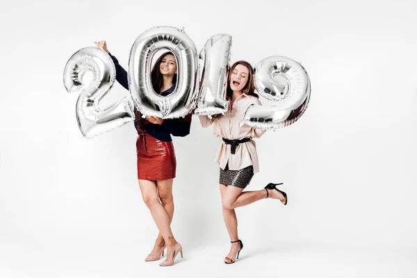 Dos hermosas chicas vestidas con una elegante ropa inteligente están sosteniendo globos en forma de números 2019 sobre un fondo blanco en el estudio — Foto de Stock