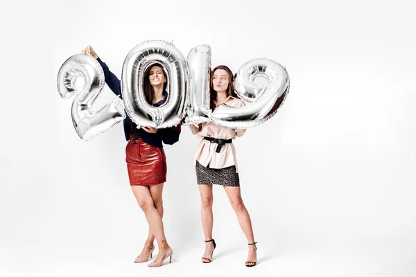 Dvě krásné dívky oblečené v stylovém elegantním oblečení drží balónky ve tvaru čísla 2019 na bílém pozadí v ateliéru — Stock fotografie