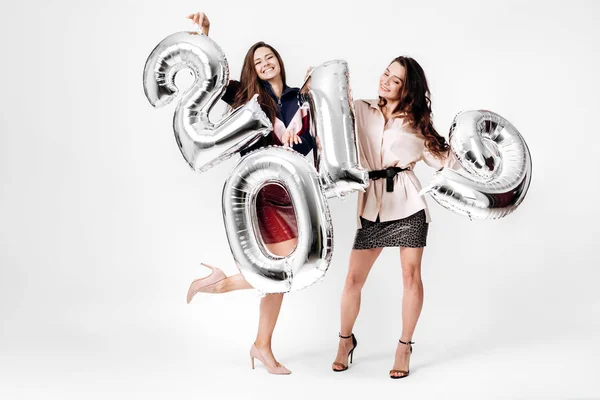 Dvě půvabné dívky oblečené v stylovém elegantním oblečení drží balónky ve tvaru čísla 2019 na bílém pozadí v ateliéru — Stock fotografie