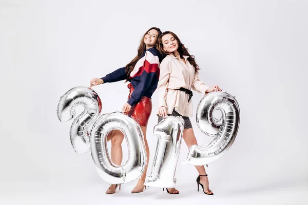 Dvě půvabné dívky oblečené v stylovém elegantním oblečení drží balónky ve tvaru čísla 2019 na bílém pozadí v ateliéru — Stock fotografie