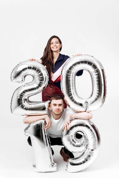 Усміхнена дівчина і хлопець, одягнені в стильний одяг, розважаються з повітряними кулями у формі цифр 2019 року на білому тлі в студії — стокове фото