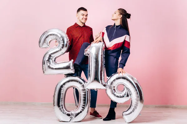 Amigos engraçados menina e cara vestido com roupas elegantes estão segurando balões na forma de números 2019 em um fundo rosa no estúdio — Fotografia de Stock