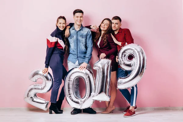 Alegre grupo de amigos de dos chicas y dos chicos vestidos con ropa elegante están sosteniendo globos en forma de números 2019 sobre un fondo rosa en el estudio — Foto de Stock