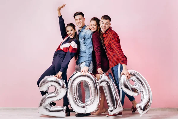 Feliz compañía de dos chicas y dos chicos vestidos con ropa elegante están sosteniendo globos en forma de números 2019 sobre un fondo rosa en el estudio — Foto de Stock