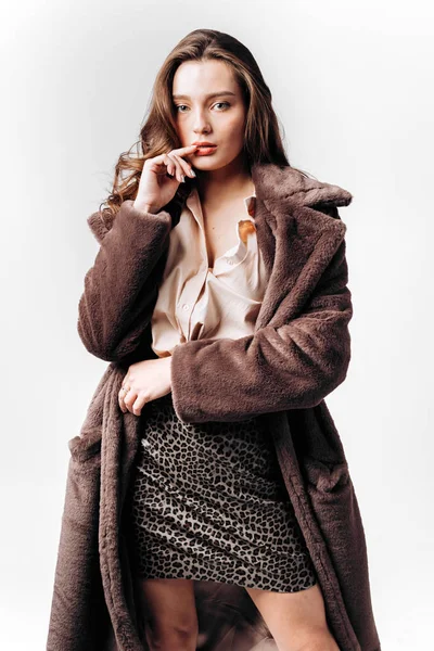 Beautiful girl in a shirt, leopard skirt and fur coat stands on the white background in the studio — Stock Photo, Image