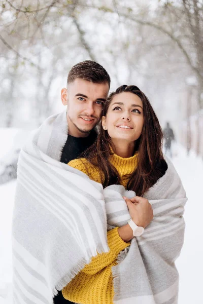 Menina de cabelos escuros encantadora vestida com uma camisola amarela e seu namorado envolto em um cachecol cinza estão de pé em uma rua nevada em um dia de inverno — Fotografia de Stock