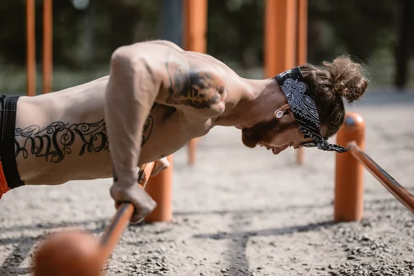 Jeune homme athlétique au bandeau avec un torse nu avec des tatouages vêtus de leggings noirs et des shorts rouges faisant des pompes sur les barres inégales à l'extérieur par une journée ensoleillée — Photo