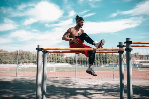 Athletic man in headband with naked torso dressed in black leggings and red shorts doing exercises on the horizontal bar on the sports ground outside