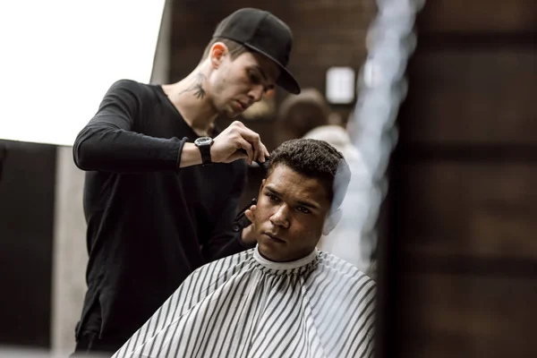 The barber dressed in black clothes  makes a razor cut hair back and sides for a stylish man sitting in the armchair in a barbershop