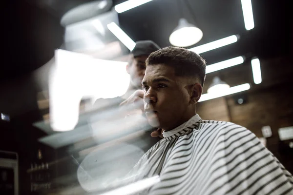 Der Modefriseur in schwarzer Kleidung schneidet einem eleganten schwarzhaarigen Mann, der in einem stilvollen Friseurladen im Sessel sitzt, die Haare messerscharf — Stockfoto