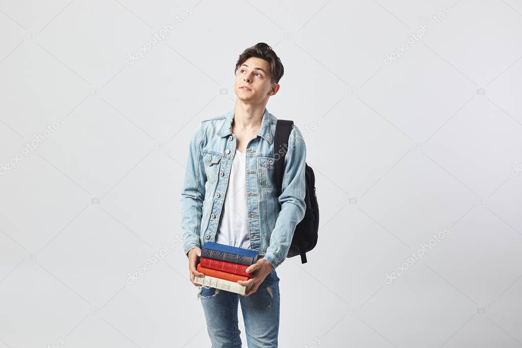Young dark-haired guy with black backpack  on his shoulder dressed in a white t-shirt, jeans and a denim jacket holds books in his hands on the white background  in the studio
