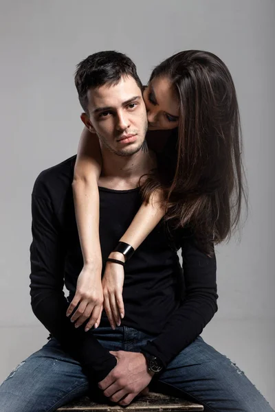 Beautiful brunette girl hugging from behind a guy dressed in a black long sleeve t-shirt and jeans sitting on a wooden chair. Young and cocky lovers — Stock Photo, Image
