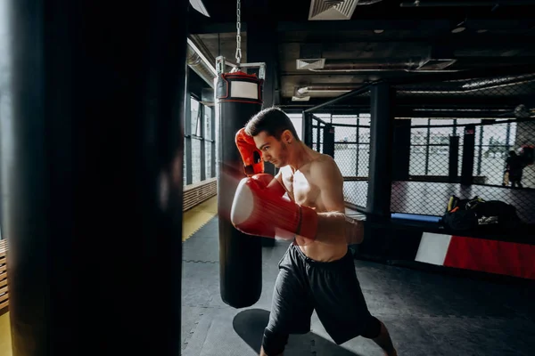 Le gars en gants de boxe rouges avec un torse nu habillé dans le short noir frappe sac de boxe dans la salle de gym — Photo